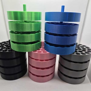 May include: A collection of 3D-printed organizers in various colors, including green, blue, black, pink, and gray. Each organizer has a cylindrical base with multiple holes and a series of stacked, circular trays. A small peg extends from the top of each organizer.