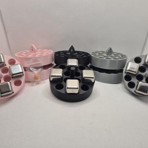 May include: Three round ice cube trays with lids, one pink, one black, and one gray. Each tray has a circular center with a small metal ice cube in each of the surrounding slots. The trays are filled with metal ice cubes.