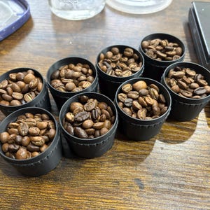 May include: Seven small black containers filled with roasted coffee beans. The beans vary in colour from light brown to dark brown. A digital scale is visible in the background, suggesting a focus on precision in coffee preparation.