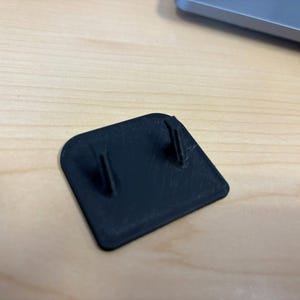 May include: A small, black, square plastic component with two vertical posts. It is positioned on a light-coloured wooden surface. The component appears to be part of a larger device or product.