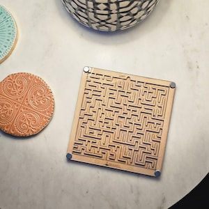 May include: A wooden maze game with a complex path design. The square game has small magnets on each corner. A round, embossed coaster and a decorative bowl are also visible.
