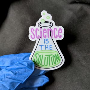 May include: A sticker featuring a science beaker design with the words "Science is the Solution." The beaker is white with a green liquid inside and the text is in pink and green. The sticker is held by a hand wearing a blue glove.