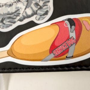 May include: A sticker featuring a cartoon illustration of a service dog inside a corn dog. The dog wears a red vest with the words "SERVICE DOG" on it. The corn dog is golden brown with a wooden stick.