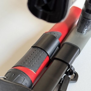 Puede incluir: Primer plano de dos postes telescópicos negros y rojos con una correa negra. Los postes están conectados entre sí con un clip de plástico negro.