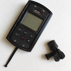 Può includere: Unità di controllo del metal detector nera con la scritta "DEUS II" e un connettore nero separato. L'unità di controllo ha uno schermo e diversi pulsanti. Un cavo nero si estende dall'unità. Gli oggetti sono su una superficie bianca.