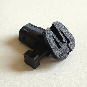 Puede incluir: Dos piezas de plástico negro, una con una superficie texturizada y un diseño en relieve, y la otra con una forma más compleja y un pequeño agujero.