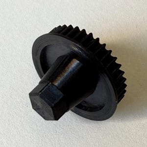 May include: A black gear with a toothed outer edge and a hexagonal base. The gear has a circular disc shape and a cylindrical post. The object is made of a solid, dark material.