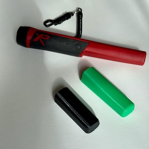 Puede incluir: Un detector de metales rojo y negro con una funda de transporte negra y una funda de transporte verde. El detector de metales tiene una correa negra y un mango negro y rojo. El detector de metales está etiquetado como "MI-6".