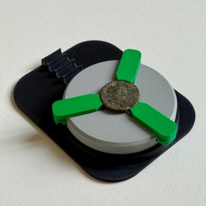 Puede incluir: Un objeto impreso en 3D, gris y negro, con tres brazos verde brillante que sostienen una moneda antigua. La moneda está centrada en un disco gris, y el conjunto se asienta sobre una base cuadrada negra.