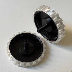 Puede incluir: Dos ruedas de plástico negro y transparente con un borde exterior dentado. Las ruedas tienen un cubo central y están diseñadas para una aplicación mecánica. El borde transparente tiene un diseño texturizado y acanalado. Las ruedas están sobre un fondo blanco.
