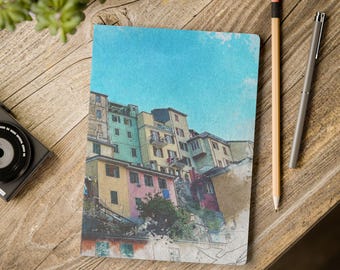 May include: A notebook with a watercolor painting of colorful buildings under a blue sky. A pencil and pen are next to the notebook on a wooden surface. A camera and plant are also visible.