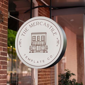 May include: A round sign for "The Mercantile Template Co." hangs outside a brick building. The sign is white with a brown border and features a line drawing of a building. The sign is suspended by a black metal bracket.