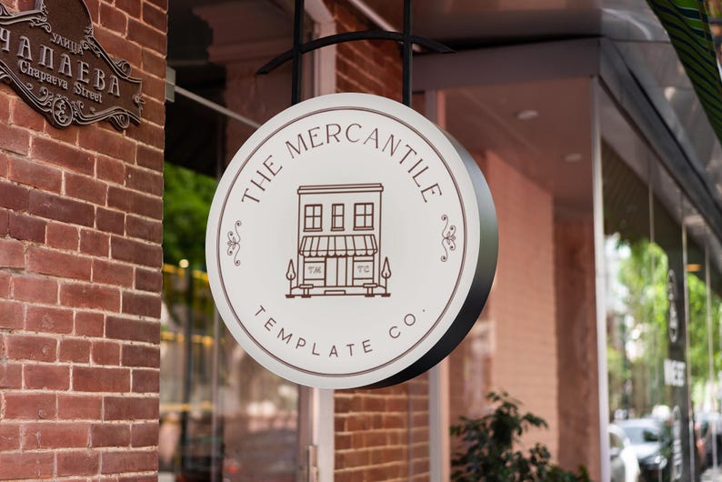 May include: A round sign for "The Mercantile Template Co." hangs outside a brick building. The sign is white with a brown border and features a line drawing of a building. The sign is suspended by a black metal bracket.