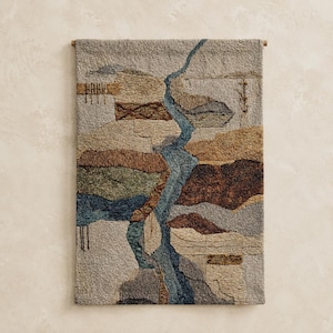 Könnte beinhalten: Ein rechteckiger Wandbehang mit einem strukturierten, getufteten abstrakten Landschaftsmotiv. Die Komposition umfasst erdige Töne in Beige, Braun, Grün und Dunkelblau, mit einem markanten zentralen blauen Element, das an einen Fluss erinnert.
