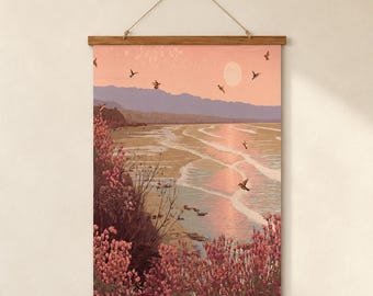 May include: A vertical wall hanging featuring a landscape print of a beach at sunset with pink and purple hues. The scene includes blooming pink flowers in the foreground, a calm ocean with gentle waves, distant mountains, and several birds.