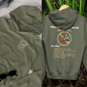 May include: A green hooded sweatshirt with the text "SERVICE DOG WORK (DO NOT PET)" on the back. The front of the sweatshirt has the text "SERVICE DOG HANDLER" with a small illustration of a dog.