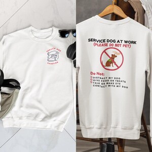 May include: White sweatshirt with a service dog logo on the front and a service dog at work sign on the back. The sign says "Service Dog at Work (Please Do Not Pet)" and includes a list of things not to do: "Do Not: Distract my dog, Give food or treats, Talk or make eye contact with my dog."