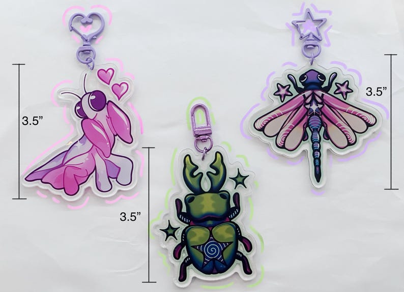 May include: Three 3.5 inch acrylic charms featuring a pink mantis, a green stag beetle, and a purple dragonfly. Each charm has a silver metal ring and a purple plastic clip.