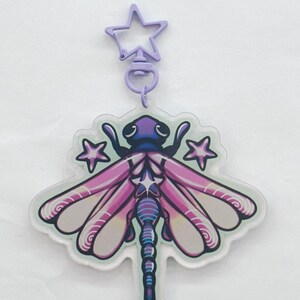 A purple, blue, and yellow colored acrylic dragonfly keychain with a plastic purple star chain surrounded by purple stars.