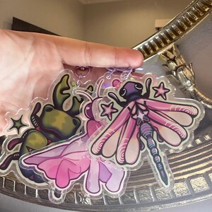 May include: Two clear acrylic charms featuring a green frog with a pink flower and a pink dragonfly with iridescent wings. Both charms have glitter accents and are attached to a silver chain.