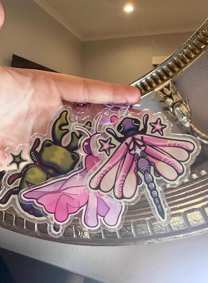 May include: Two clear acrylic charms featuring a green frog with a pink flower and a pink dragonfly with iridescent wings. Both charms have glitter accents and are attached to a silver chain.