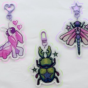 May include: Three colorful keychains featuring a pink praying mantis, a green stag beetle, and a purple dragonfly. Each keychain has a silver clip and a star charm.
