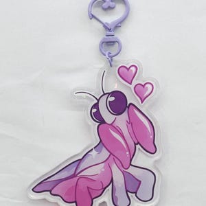A purple and pink colored acrylic orchid mantis keychain with a plastic purple heart chain surrounded by pink hearts.