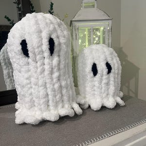 May include: Two white, plush ghost decorations with black oval eyes. One ghost is larger than the other. The ghosts are on a grey surface. A white lantern with lights is in the background.