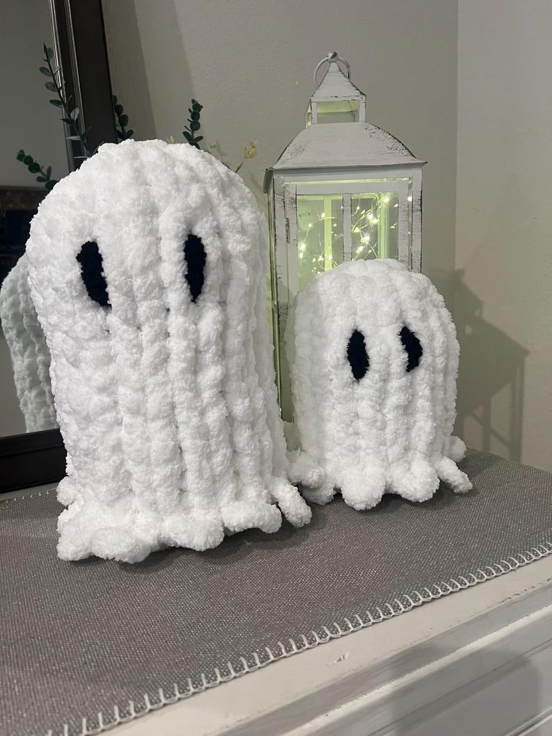 May include: Two white, plush ghost decorations with black oval eyes. One ghost is larger than the other. The ghosts are on a grey surface. A white lantern with lights is in the background.