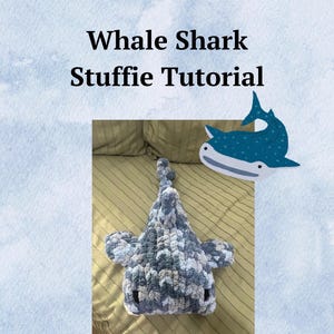 May include: A grey and white crocheted whale shark stuffed animal on a striped bedspread. The stuffed animal has a cartoon whale shark illustration above it.