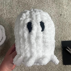May include: A handmade, white, plush ghost with black eye cutouts. The ghost has a fluffy texture and a scalloped bottom edge. The ghost is a Halloween or spooky season decoration.
