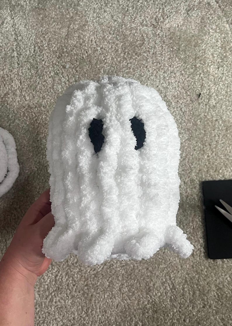 May include: A handmade, white, plush ghost with black eye cutouts. The ghost has a fluffy texture and a scalloped bottom edge. The ghost is a Halloween or spooky season decoration.