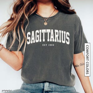 May include: Dark grey t-shirt with the word "SAGITTARIUS" in white block letters. Below the word is "EST 1993". The shirt is a Comfort Colors brand.