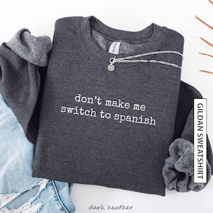 May include: Dark grey sweatshirt featuring the white text "don't make me switch to spanish". A silver necklace with a sunburst pendant is draped across the front. The sweatshirt is folded over denim shorts.