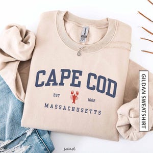 Puede incluir: Sudadera color arena con "CAPE COD" en letras azul marino, un gráfico de langosta roja y "EST 1602 MASSACHUSETTS" impreso debajo. Un collar plateado con un colgante de sol descansa sobre la sudadera. La sudadera está doblada sobre unos vaqueros azules.