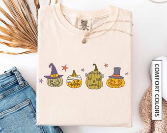 Puede incluir: Camiseta de color marfil con cuatro diseños de calabazas de Halloween. Las calabazas son verdes, naranjas y amarillas, cada una con un sombrero o envoltura diferente. La camiseta tiene las palabras "COMFORT COLORS" impresas en el lateral. La palabra "ivory" está impresa en la parte inferior.