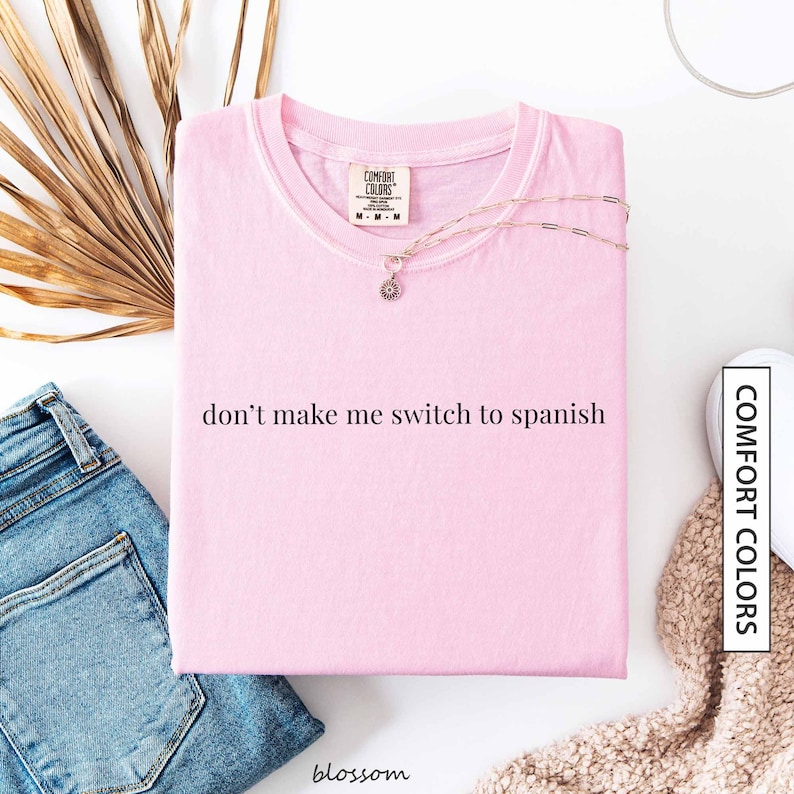 a pink t-shirt with the text "don't make me switch to spanish" printed on it, along with a necklace and some other clothing items.