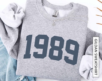 May include: A sport grey crewneck sweatshirt featuring the dark navy blue text 1989 printed across the chest. The garment has a ribbed collar, cuffs, and hem, with a Gildan brand label visible at the inner neckline. A silver chain necklace rests on top.