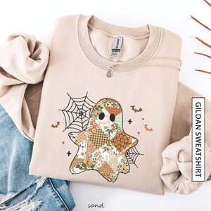 Op de afbeelding: Zandkleurig Gildan sweatshirt met een patchwork spookontwerp met bloemen- en geometrische patronen. Het ontwerp bevat een spinnenweb, vleermuizen en kruisen. Een zilveren ketting is over het sweatshirt gedrapeerd. Het woord "sand" staat onderaan.