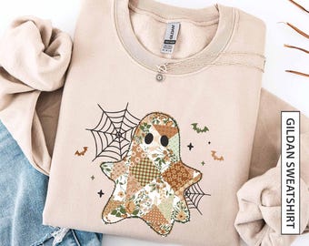 Pode incluir: Camisola Gildan cor areia com um design de fantasma de patchwork com padrões florais e geométricos. O design inclui uma teia de aranha, morcegos e cruzes. Um colar de prata está sobre a camisola. A palavra "sand" está na parte inferior.
