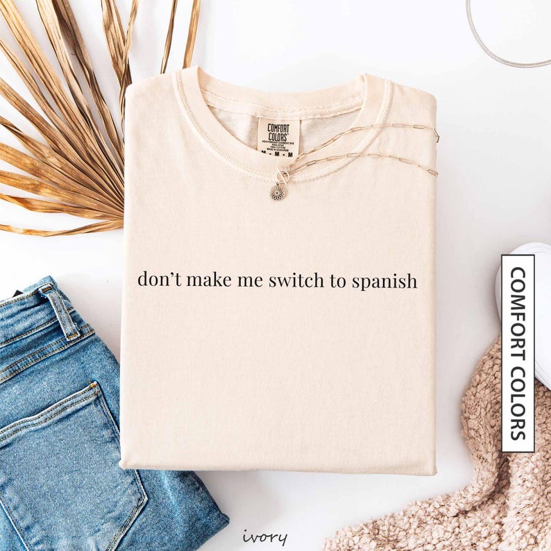 a white t-shirt with the text "don't make me switch to spanish" printed on it, along with a pair of blue jeans and a beige sweater.