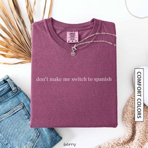 a purple t-shirt with the text "don't make me switch to spanish" printed on it, along with a necklace and some other clothing items.