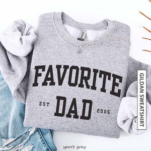 May include: A sport grey sweatshirt with the words "FAVORITE DAD" in bold black letters. Below the text are the words "EST" and "DAD" with the year "2005". A silver necklace with a pendant is draped across the sweatshirt.