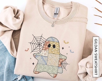 May include: Sand-coloured Gildan sweatshirt with a patchwork ghost design featuring floral and geometric patterns. The ghost is surrounded by a spiderweb, bats, and stars. A silver necklace with a sun pendant is on the sweatshirt. The text "Gildan Sweatshirt" is on the right.