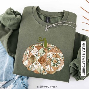 May include: Military green Gildan sweatshirt with a patchwork pumpkin design. The pumpkin is made up of different fabric patterns in brown, cream, and green. A silver necklace with a small pendant is draped on the sweatshirt. The words "Gildan Sweatshirt" are printed vertically on the right side.