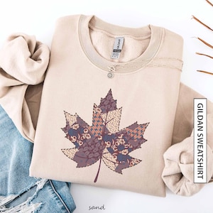 May include: A sand-coloured Gildan sweatshirt with a large maple leaf design. The leaf is made of patterned fabric patches in purple, brown, and blue. A silver necklace with a small pendant rests on the sweatshirt. The word "sand" is at the bottom.