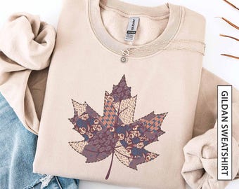 May include: Sand-colored Gildan sweatshirt featuring a large, detailed maple leaf design. The leaf is composed of various patterned fabric patches in shades of purple, brown, and blue. A delicate silver necklace with a small pendant rests on the sweatshirt. The word "sand" is at the bottom.