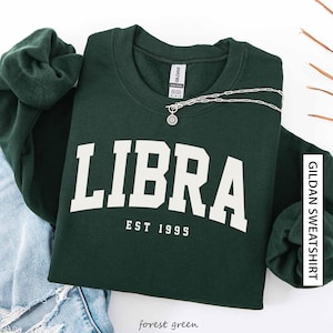 Könnte beinhalten: Forstgrünes Sweatshirt mit dem Wort "LIBRA" in weißen Blockbuchstaben und "EST 1995" darunter. Eine silberne Halskette mit Sonnenanhänger ist über dem Sweatshirt drapiert. Das Sweatshirt ist mit einer Jeans gefaltet.