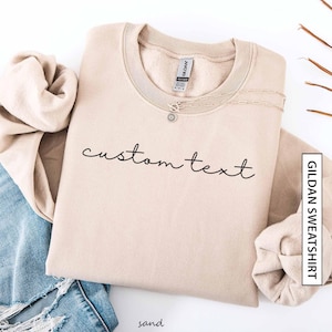 May include: A sand-colored Gildan sweatshirt with the words "custom text" in black script. A silver necklace with a small pendant rests on the sweatshirt. The sweatshirt is folded with a pair of blue jeans and a rolled-up beige sleeve.