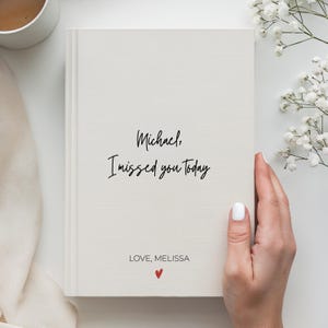May include: A cream-coloured hardback book featuring the black text "Michael, I missed you Today" printed in the centre. Below this, the text "LOVE, MELISSA" appears in a smaller font, followed by a small, centred red heart icon at the bottom.